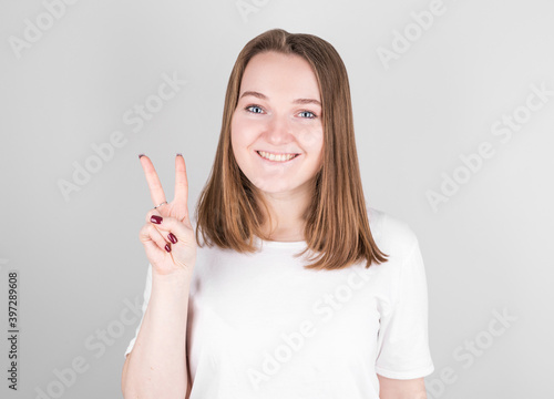Young smiling woman stands in a white T-shirt on a gray background and shows a peace sign.