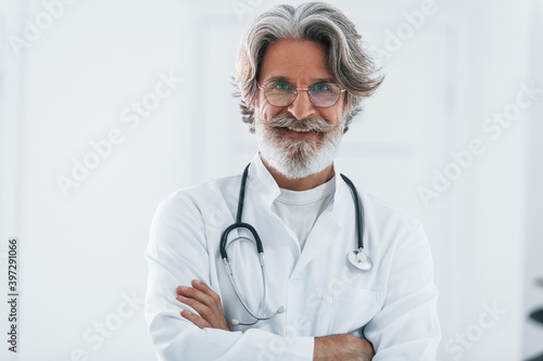 Portrait of senior male doctor with grey hair and beard in white coat standing indoors in clinic