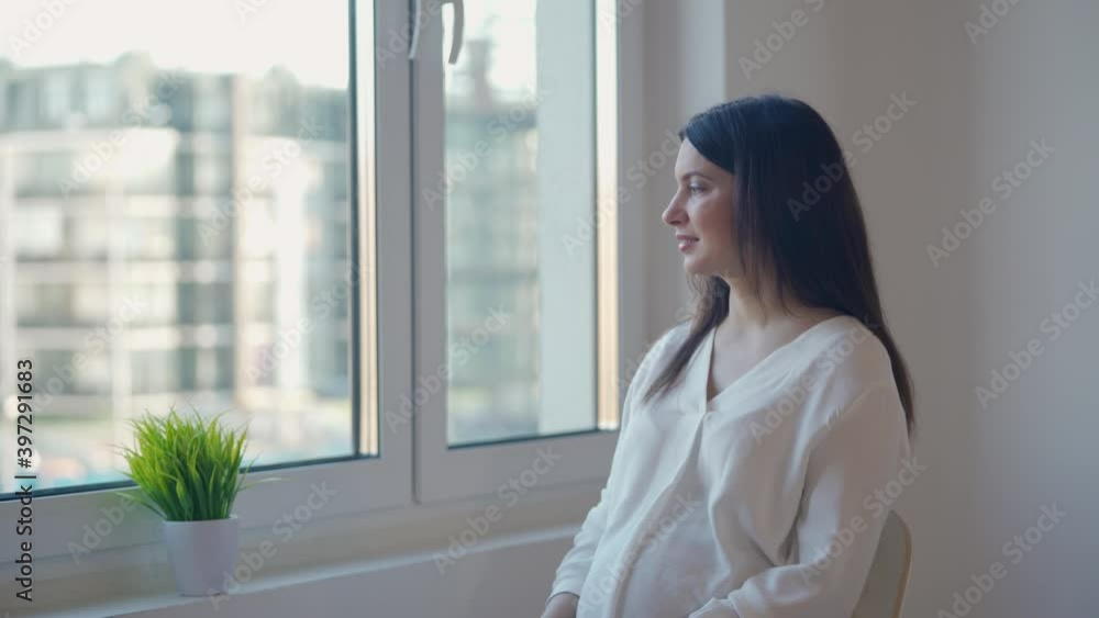 Beautiful pregnant woman by the window in new flat looking at outside, brunette lady posing in white apartment, moving to new house, home construction.