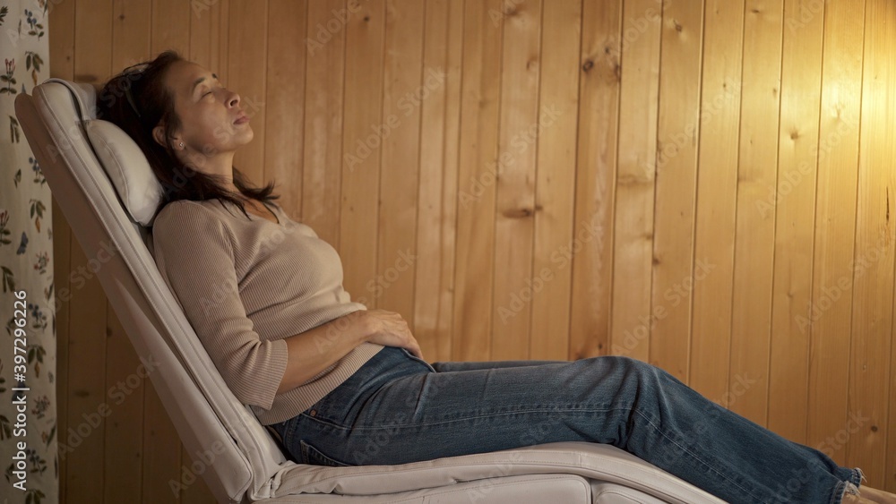 Asian woman resting sleeping in the armchair. Profile shot of female lying on the long armchair with eyes closed, evening light on background of wooden panels Asian woman resting sleeping in the armchair. Profile shot of female lying on the long armchair with eyes closed, evening light on background of wooden panels