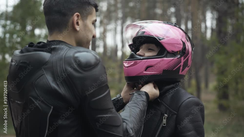 Portrait of young Caucasian girlfriend smiling to boyfriend as Middle Eastern man fastening her pink motorcycle helmet. Loving interracial happy couple of motorbikers preparing for a ride outdoors. Portrait of young Caucasian girlfriend smiling to boyfriend as Middle Eastern man fastening her pink motorcycle helmet. Loving interracial happy couple of motorbikers preparing for a ride outdoors.