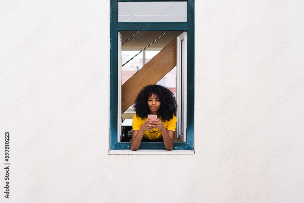 © David Prado/Stocksy - Cheerful ethnic woman using phone while looking out window © David Prado/Stocksy - Cheerful ethnic woman using phone while looking out window