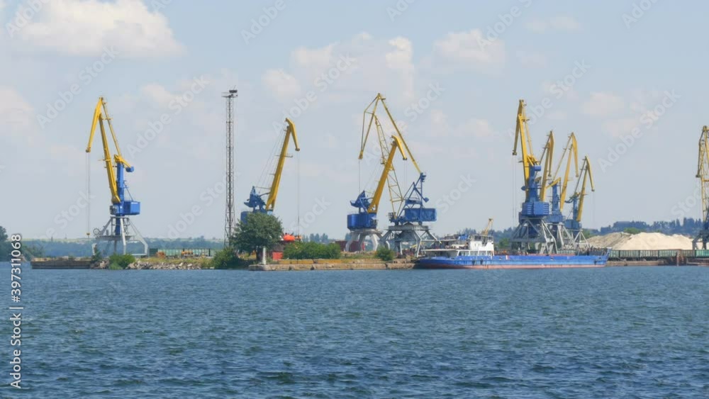 A row of construction or cargo cranes on the banks of a river or pond against the background of a barge standing nearby A row of construction or cargo cranes on the banks of a river or pond against the background of a barge standing nearby