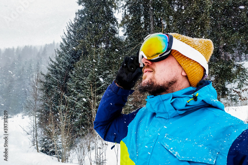 Close up portrait of man skier in hat and ski goggles mask looking at mountain. it's snowing