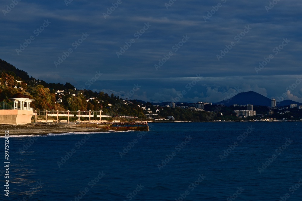 Seascape with a jutting cape with a rotunda in the rays of sunset against the background of mountains and the city