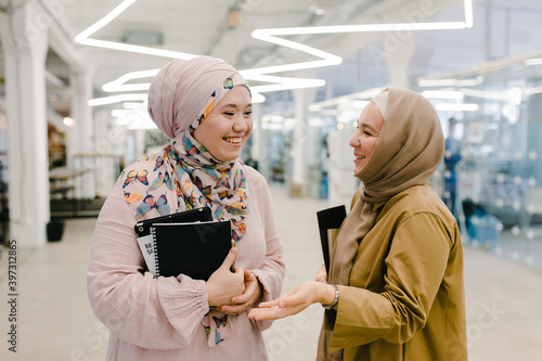 Happy Muslim female colleagues with copybooks chatting in hallway photo