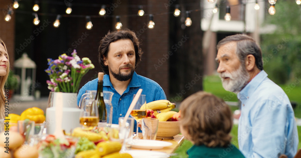 Obraz premium Caucasian family sitting at table in garden at back yard. Old grandfather telling story to grandchild and son with wife. Senior man talking with adults children and small grandson. Weekend spending.
