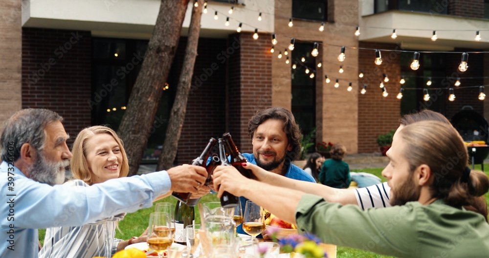Obraz premium Smiled cheerful Caucasian family laughing, talking and toasting at barbecue outdoor on weekend day. Happy people of different ages saying toasts and cheering with beer bottles. Drinks in hands.