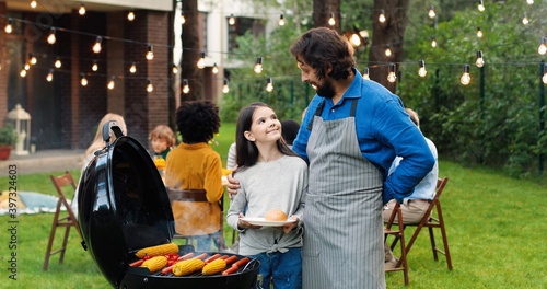 Portrait of Caucasian happy father and little daughter standing at back yard at barbecue, hugging, smiling and cooking corn and meat. Dad with small pretty girl preparing food for lunch on fresh air.