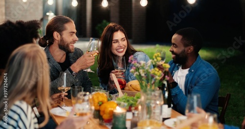 Multi ethnic joyful young friends talking, laughing and toasting with drinks at party table in evening. Mixed-races women and men rising glasses with toasts at night and drinking alcohol. Celebrating.