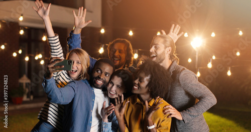 Mixed-races young happy and cheerful friends, men and women smiling and posing to smartphone while taking selfie photo at night. late evening party. Multi ethnic men and women having fun.