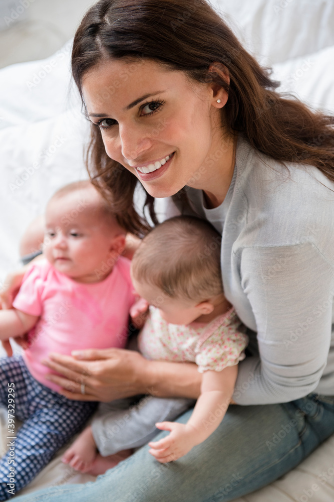 Caucasian mother sitting on bed holding twin baby daughters Caucasian mother sitting on bed holding twin baby daughters