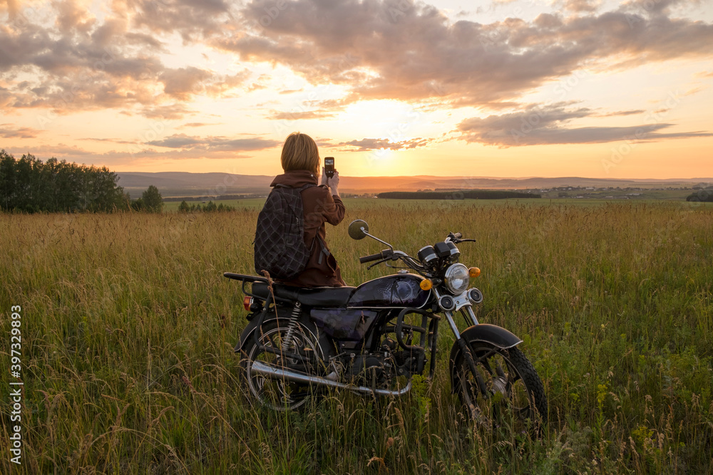Caucasian woman with motorcycle in field photographing sunset Caucasian woman with motorcycle in field photographing sunset