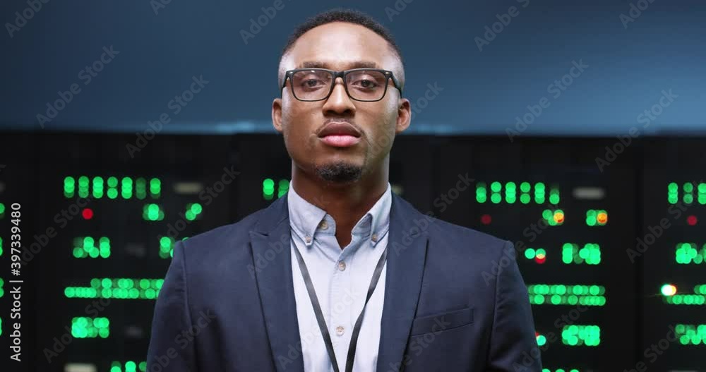 Close up portrait of African American handsome male system administrator in suit stand in big data center with server racks looking at camera Man worker in glasses alone in server room Digital concept Close up portrait of African American handsome male system administrator in suit stand in big data center with server racks looking at camera Man worker in glasses alone in server room Digital concept
