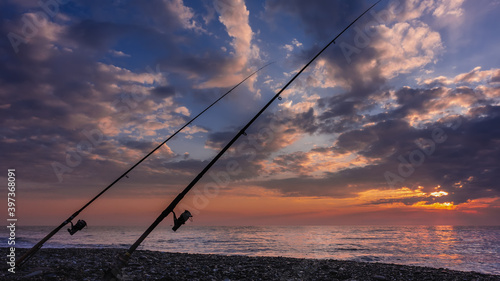 Sea fishing at sunset. Two fishing rods stand obliquely on a pebble beach. The clouds in the blue sky are tinted pink, orange, lilac. Sunny path on the water. Russia. Sochi
