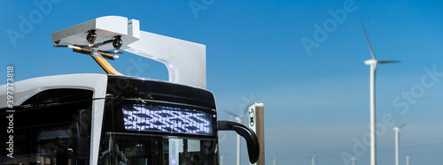 Electric bus at a stop is charged by pantograph on the background of wind turbines 