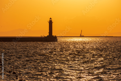 夕日に照らされて、キラキラ光る海面と灯台の元の防波堤で釣りをする人々