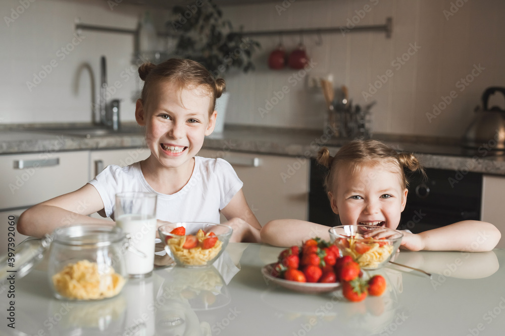 Happy having fun Little siblings girls having breakfast of  with milk and ripe strawberries in a real kitchen interior. Healthy breakfast in the morning with family