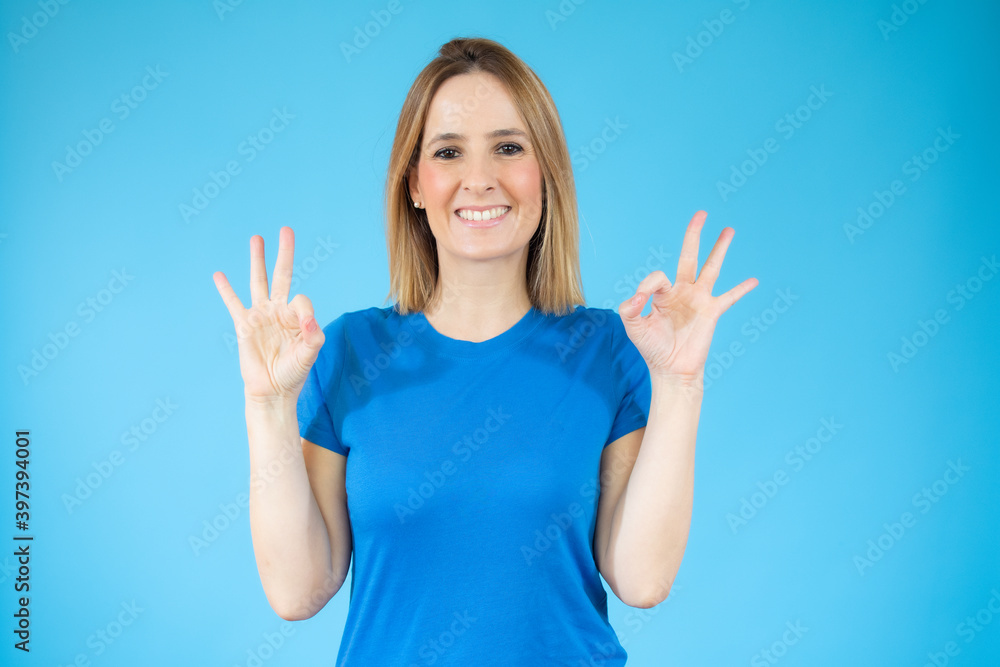 Fototapeta premium Young beautiful woman in casual t-shirt standing over blue isolated background smiling positive doing okay sign with hand and fingers. Successful expression.