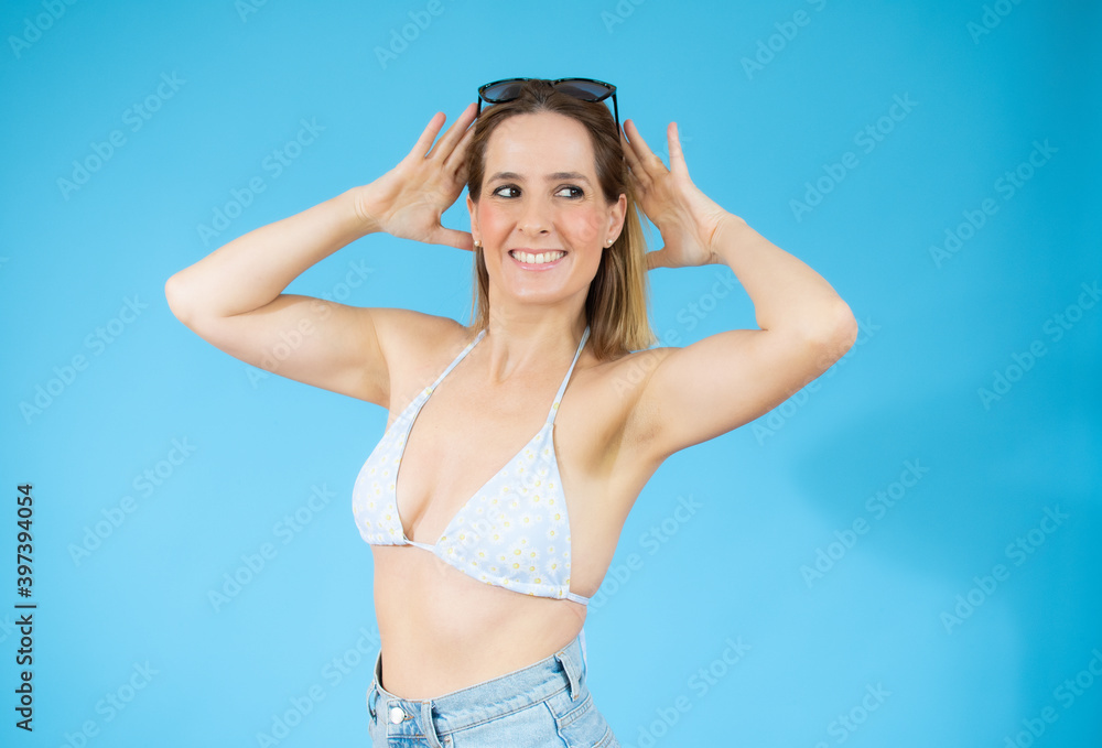 Fototapeta premium Young beautiful woman wearing bikini over isolated blue background looking away to side with smile on face, natural expression. Laughing confident.