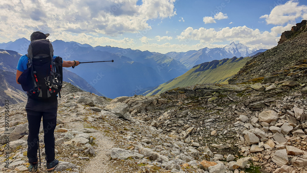 Obraz premium A man pointing with hiking stick in direction if Gro?glockner, while hiking in Heiligenblut region in Austria. There are many high chains of Alps behind him. Narrow pathway. Looking for directions