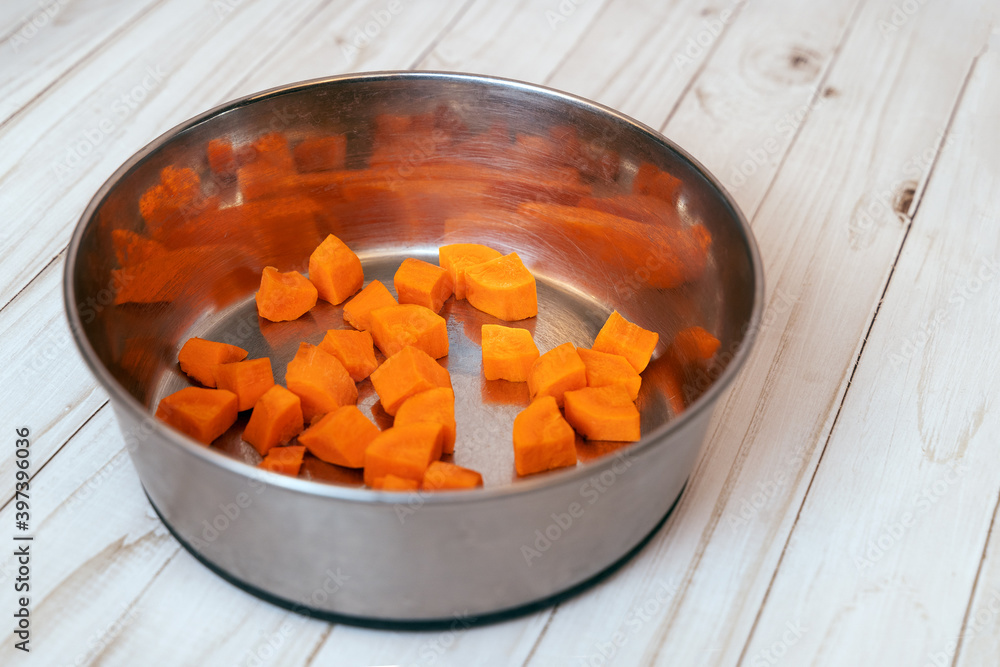 Metal pet bowl with cut carrot inside. First step of natural organic daily meal ration preparation, BARF diet for dogs concept. Copy space, white wooden table background. Metal pet bowl with cut carrot inside. First step of natural organic daily meal ration preparation, BARF diet for dogs concept. Copy space, white wooden table background.