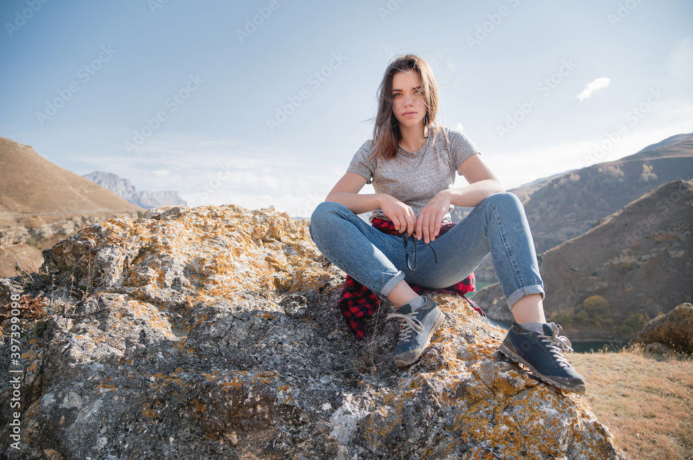 Attractive brooding young caucasian woman in gray T-shirt and jeans sits on a cliff against the mountains on a sunny day