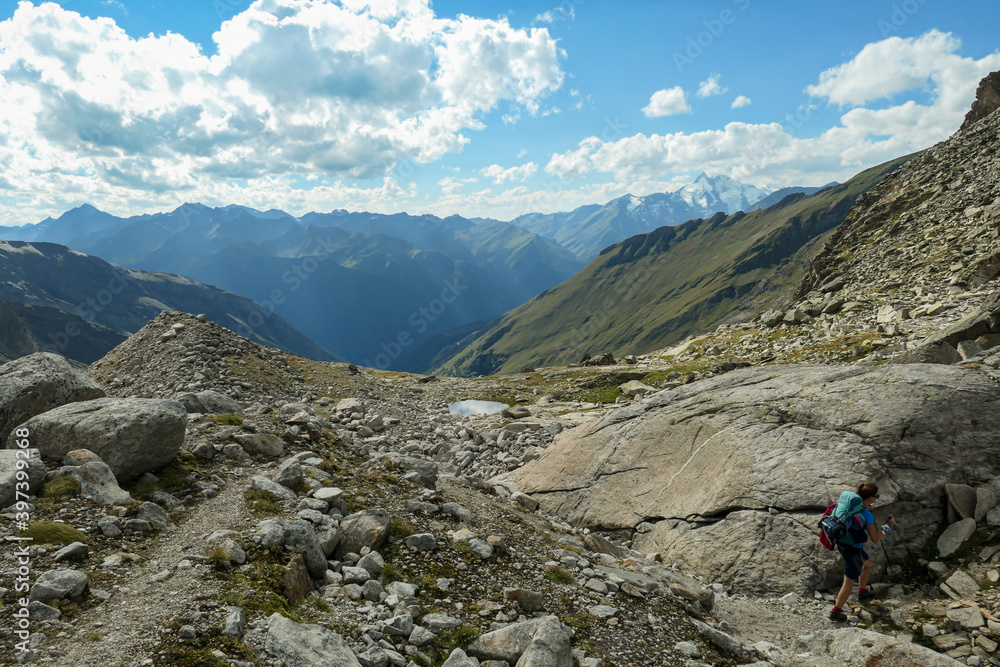 Panoramic view from Hohe Sonnblick in Austrian Alps on Gro?glockner.  A woman with hiking backpack hiking through a narrow pathway through the stony landscape, full of lose stones and boulders.