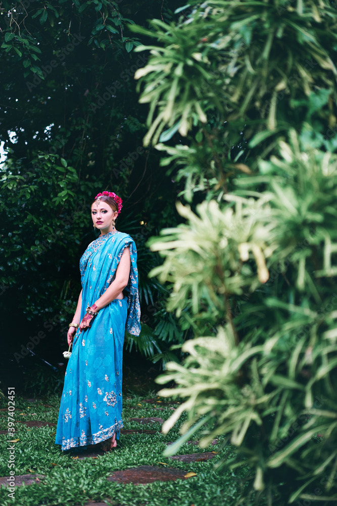 Beautiful young caucasian woman in traditional indian clothing with bridal makeup and jewelry. Bollywood dancer in Sari and henna on hands walking on green garden.
