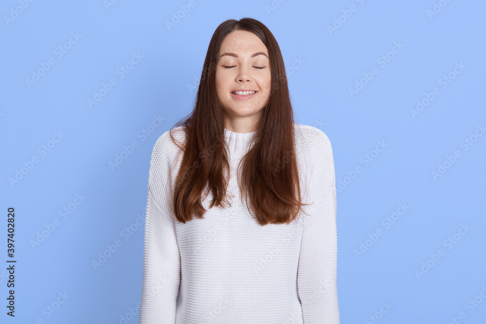 Happy dreamy woman keeps eyes closed, smiles positively, dressed in casual white sweater, isolated over blue background, wishes best, dreams about something pleasant. Happy dreamy woman keeps eyes closed, smiles positively, dressed in casual white sweater, isolated over blue background, wishes best, dreams about something pleasant.