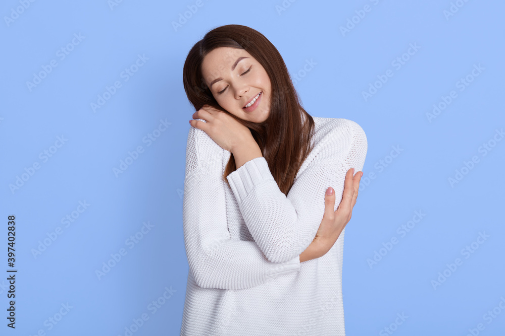 Good looking lady hugging herself, keeping eyes closed, wearing white casual jumper, posing isolated over blue background, smiling female with positive and gentle emotions. Good looking lady hugging herself, keeping eyes closed, wearing white casual jumper, posing isolated over blue background, smiling female with positive and gentle emotions.