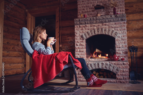 Cozy home. Pretty young woman drinking hot  beverage near the fireplace.