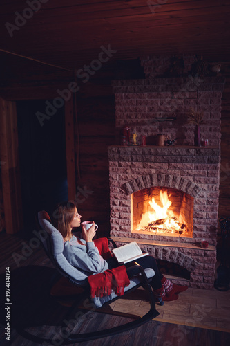 Cozy home. Pretty young woman drinking hot beverage near the fireplace.