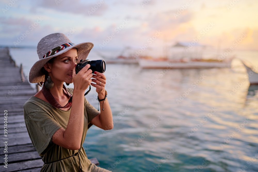 Obraz premium Photography and travel. Young woman in hat holding camera sitting on wooden fishing pier with beautiful tropical sea view.