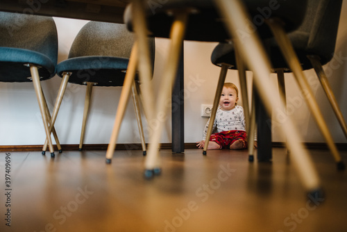 Adorable baby boy crawling underneath table. Real life toddler infant crawls under table at living-room or kitchen. Beautiful kid under the table.
