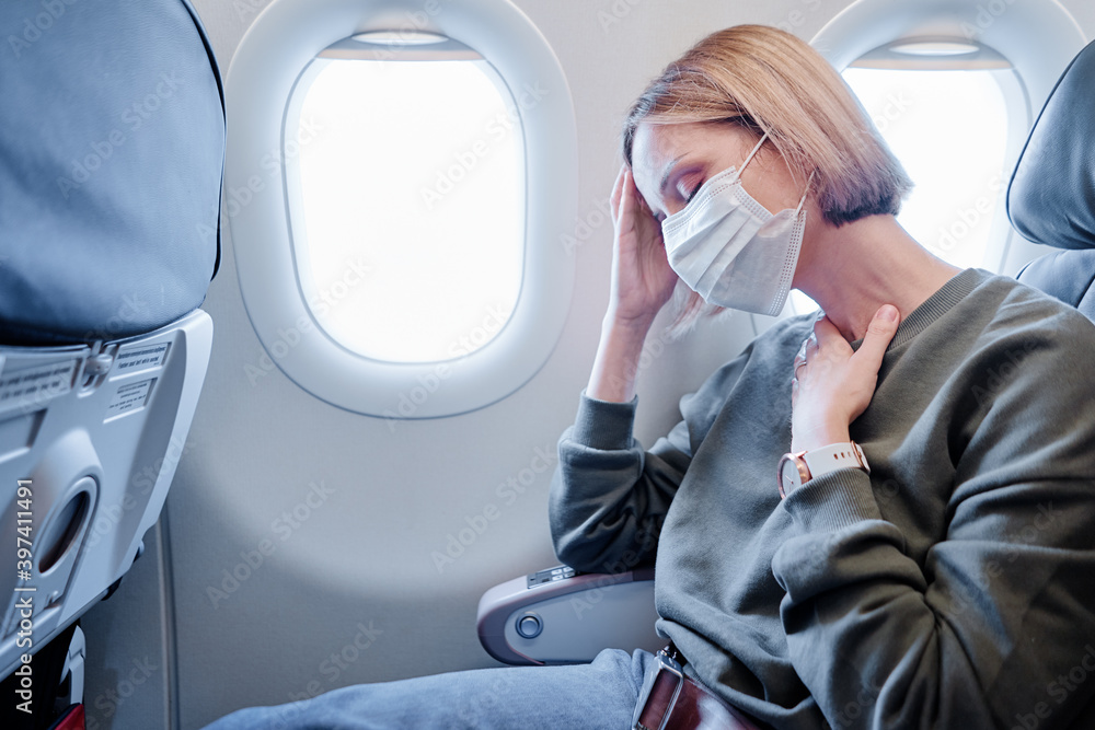 COVID-19 Young woman with face mask feeling unwell on plane. Fear of flying woman in airplane. Stress, headache, motion sickness and airsickness on plane during pandemic. - obrazy, fototapety, plakaty 