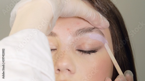 Correction of a shape of eyebrows with hot wax. Brow master applying wax on the eyebrow of female face. Wax correction of the shape of the eyebrows with spatula. Beauty industry. Close up. 