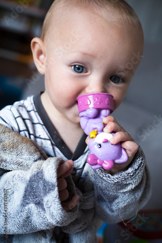 Baby girl in grey clothes and bathrobe holds in hands and nibble a violet toy cat. First tooth. Closeup portrait of blue eyecaucasian child. Teething problem. After bath. Tender and emotional concept photo