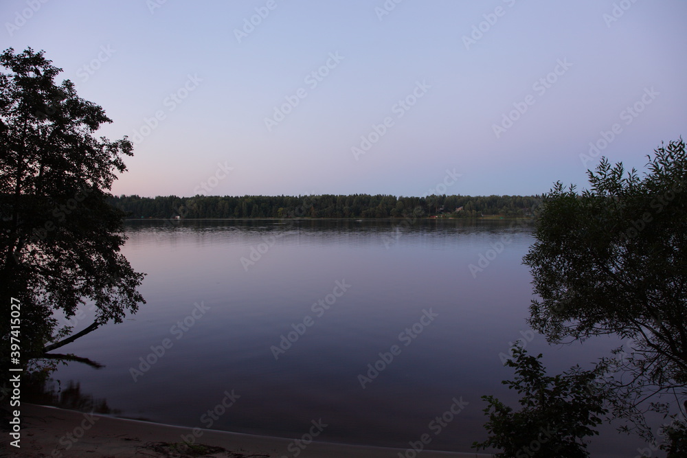 Beautiful view between the trees from riverbank on Volga river calm water, far shore with dark forest and clear blue sky at quiet summer night, Russian natural river landscape Beautiful view between the trees from riverbank on Volga river calm water, far shore with dark forest and clear blue sky at quiet summer night, Russian natural river landscape
