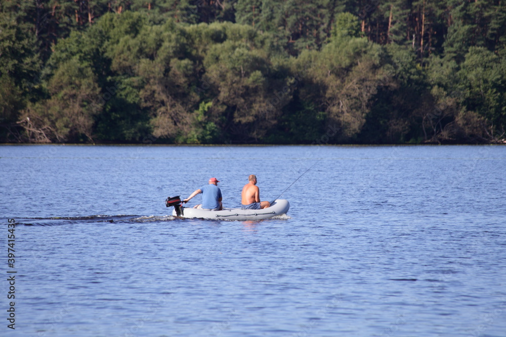 Two fishermen floating and fishing on small PVC inflatable motor boat in the middle of the river without life jackets on the far Bank with a green forest background at summer day, rest on the water