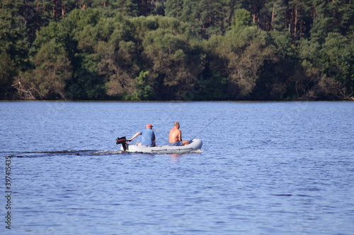 Two fishermen floating and fishing on small PVC inflatable motor boat in the middle of the river without life jackets on the far Bank with a green forest background at summer day, rest on the water