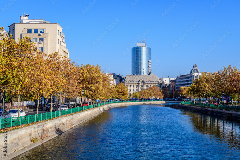 Fototapeta premium Landscape with Dambovita river, old buildings and yellow, orange and brown leaves in large trees in the center of Bucharest, Romania, in a sunny autumn day.