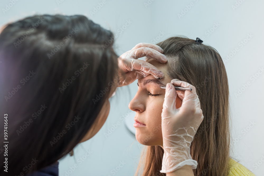 Fototapeta premium The master corrects the makeup, shapes it with a thread and plucking the eyebrows with tweezers in the beauty salon.