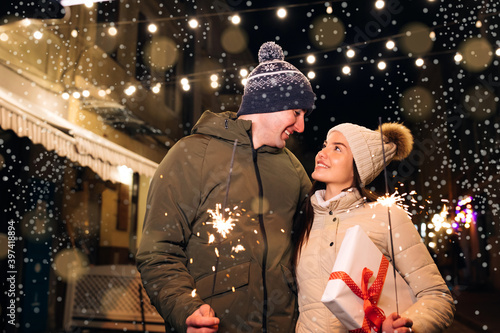 Two people celebrate New Year on the street. Close up portrait of young happy couple standing on the street hugging holding sparklers. Young couple in love holds sparklers in their hands