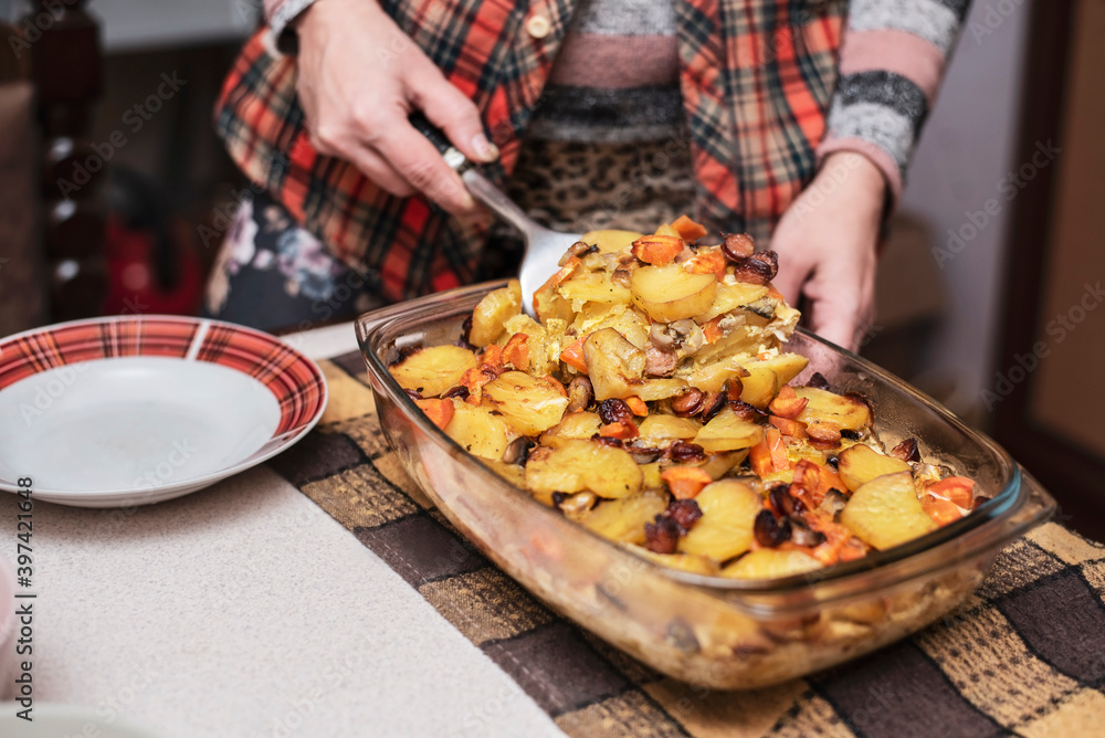 The woman cooked a delicious dinner at home - potatoes baked with vegetables, meat and sausages, and serves food to her family, for which she tries so hard and works
