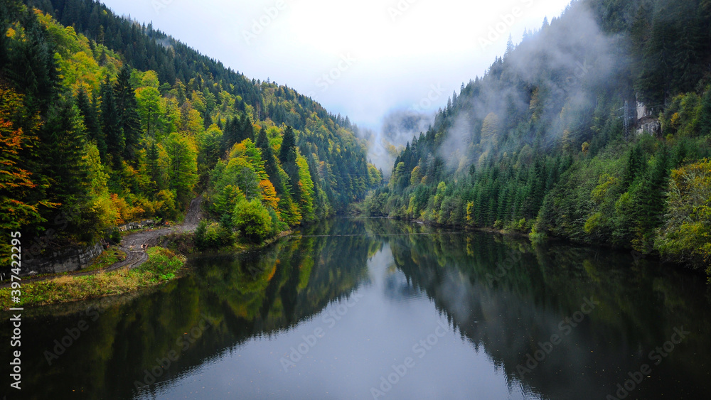 Autumn colored trees are reflecting into the water of Balindru Lake. Hazy and misty atmosphere. Fog is covering the wooded hills.