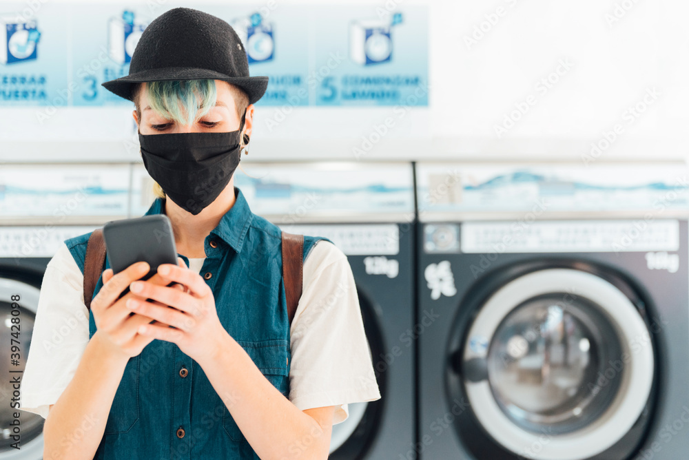 Bored young woman chatting with her friends via her smartphone while waiting for her laundry to be washed. new normal in public spaces. Bored young woman chatting with her friends via her smartphone while waiting for her laundry to be washed. new normal in public spaces.