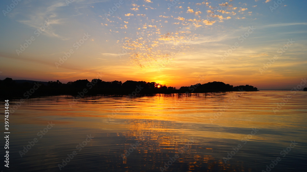 Beautiful sunset on the river. The sun sets behind the edge of the trees. The clouds are unusually illuminated by the setting sun