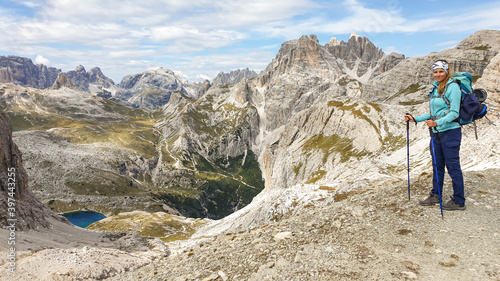 A woman with hiking backpack and sticks enjoying the view on Italian Dolomites. There are sharp and steep mountains. At the bottom of a small valley there is a small, navy blue lake. Raw landscape