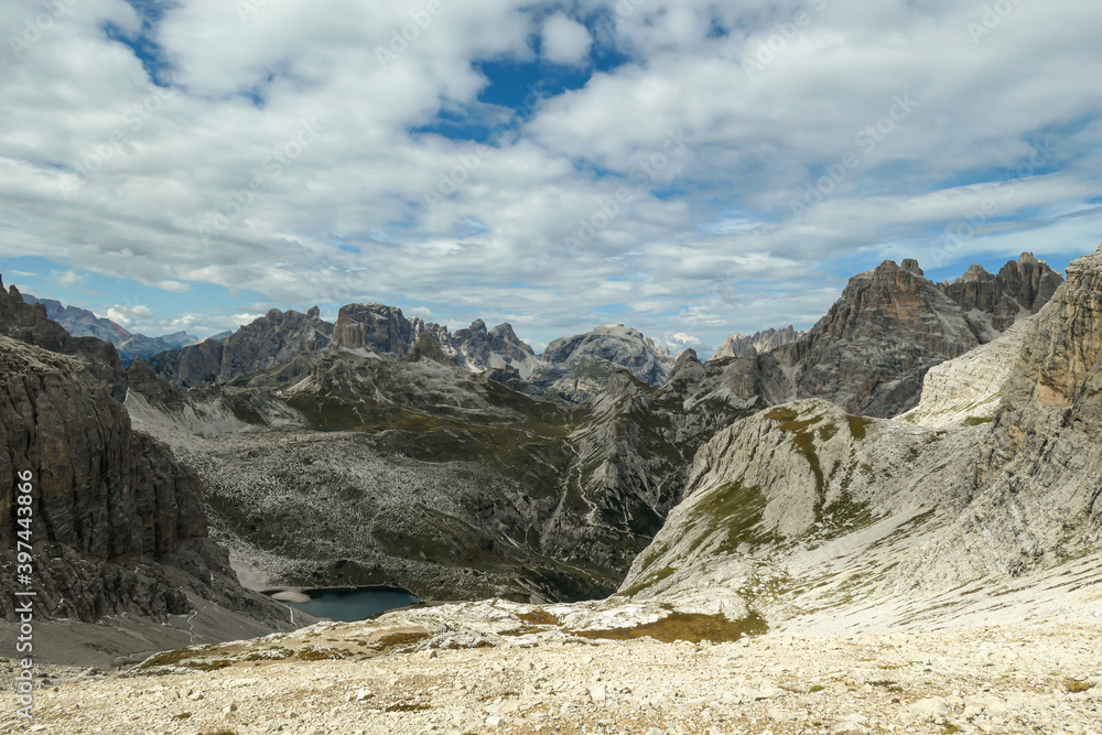 A panoramic view on Dolomites in Italy. There are sharp and steep mountain slopes around. At the bottom of a small valley there is a small navy blue lake. The sky is full of soft clouds. Raw landscape